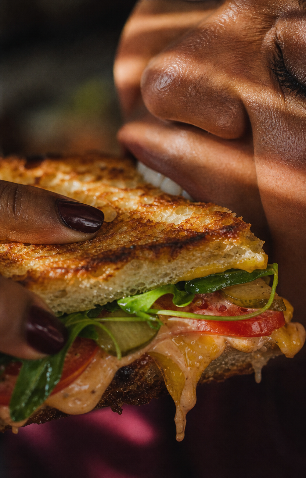 Extreme close-up of someone biting into a grilled sandwich with greens, tomato, pickles, and melted cheese.